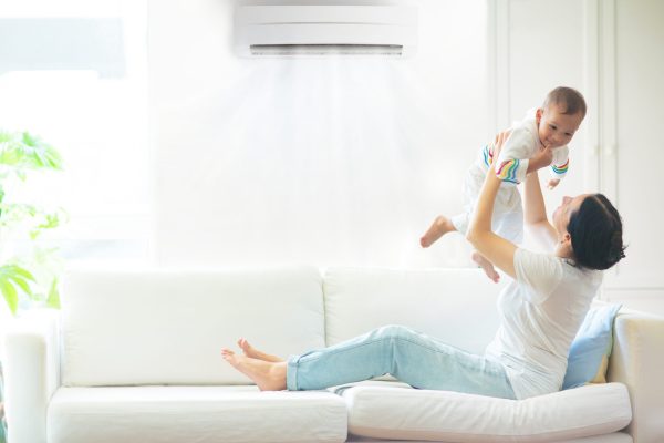 Asian mother and child with air conditioner remote control. Comfortable temperature at family home. Cooling device. Mom and baby boy on couch under cold breeze. Air conditioning on hot summer day.
